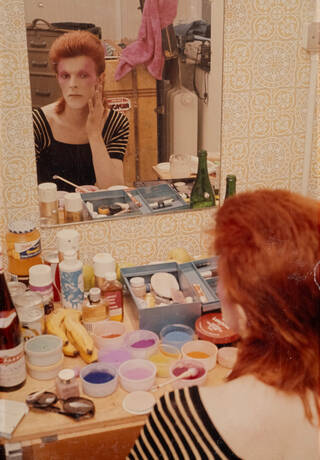 Photograph of David Bowie applying makeup as Ziggy Stardust in a mirror