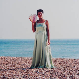 Black woman standing on a pebble beach, holding a large pink shell to her ear. She wears a green floor-length dress