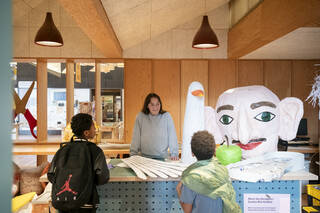 A woman wearing a blue jumper smiles at 2 children, she is surrounded by large cardboard props in the shape of a big face, scissors and a seagull